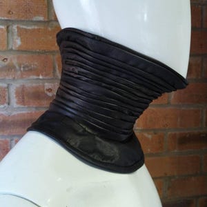 May include: A black neck warmer featuring a series of horizontal, gathered pleats that wrap around the neck. The accessory has a smooth, flat base at the bottom and a structured, cylindrical shape that fits snugly around the neck area.