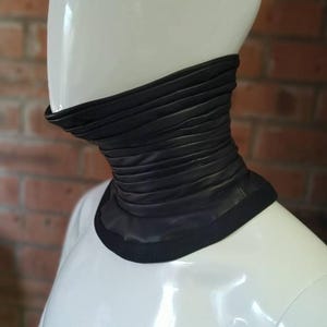 May include: A black leather neck gaiter featuring a series of horizontal, gathered pleats that create a textured, layered appearance. The accessory has a smooth, solid black trim along the top and bottom edges, fitting snugly around the neck area.