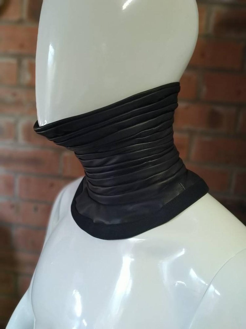 May include: A black leather neck gaiter featuring a series of horizontal, gathered pleats that create a textured, layered appearance. The accessory has a smooth, solid black trim along the top and bottom edges, fitting snugly around the neck area.