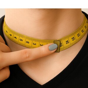 May include: A yellow flexible measuring tape wrapped around a neck, showing black numerical markings in centimeters. The tape is secured with a small rectangular metal clasp, indicating a measurement of approximately 60 centimeters in total length.