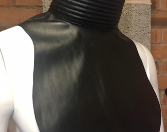 May include: A black faux leather dickey featuring a high, ribbed mock neck collar. The garment covers the upper chest area with a smooth, matte finish and a rounded hemline, designed to be worn under other clothing items for a layered appearance.