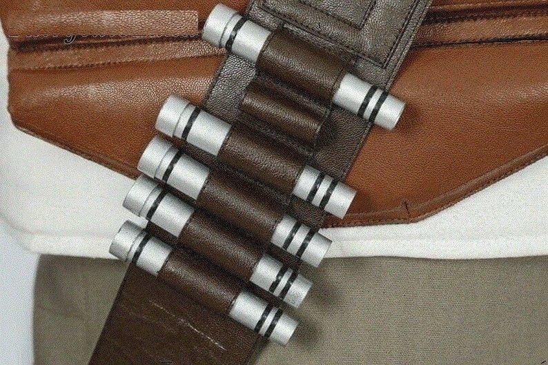 May include: Close-up of a brown leather belt with silver and black cylindrical details. The belt is attached to a brown leather garment. The design suggests a costume or cosplay accessory.