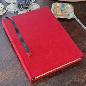 Op de afbeelding: Een rood notitieboek met een zwart lint als bladwijzer en een decoratief rood en goud accent. Het notitieboek is rechthoekig en lijkt gemaakt van een zacht, getextureerd materiaal. De pagina's zijn wit en het notitieboek is gesloten.