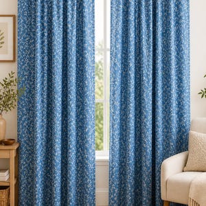 Könnte beinhalten: Zwei blaue Vorhänge mit Blumenmuster hängen vor einem Fenster. Die Vorhänge sind mittelblau mit einem weißen Blumenmuster. Ein gerahmtes Bild und ein kleiner Tisch mit einer Vase befinden sich links von den Vorhängen. Rechts befindet sich ein weißer Stuhl mit einer Decke.