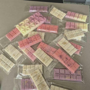 Puede incluir: Una colección de pastillas de cera rectangulares en envoltorios individuales de plástico transparente. Las barras presentan varios tonos de rosa, morado y blanco, con algunas piezas que contienen purpurina visible o motas de color.