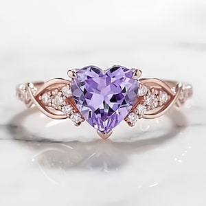 Op de afbeelding: Een roségouden ring met een centraal hartvormige paarse edelsteen. De band is aan de zijkanten bezet met kleine, heldere ronde steentjes in een ingewikkeld, verweven metaalontwerp dat naar de centrale hartvormige zetting toe loopt.