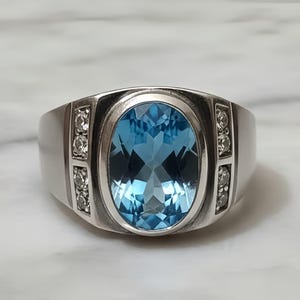 May include: A silver ring featuring a large, oval-cut, light blue gemstone as the centerpiece. The ring has a wide band and is accented with smaller, clear gemstones on each side of the main stone.