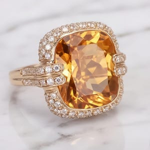 May include: A gold ring featuring a large, square-cut, amber-colored gemstone, surrounded by a halo of small, clear gemstones. Additional small gemstones adorn the ring's band and shoulders. The ring is set against a white marble background.