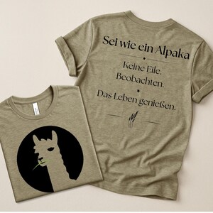 Könnte beinhalten: Ein olivgrünes T-Shirt mit einer schwarzen kreisförmigen Grafik, die die Silhouette eines hellen Alpakas zeigt, das einen grünen Grashalm kaut. Auf der Rückseite steht der schwarze Text: Sei wie ein Alpaka. Keine Eile. Beobachten. Das Leben genießen.