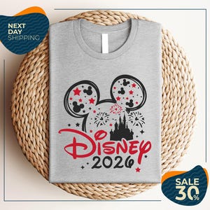 A gray t-shirt with a Disney-themed design, including Mickey Mouse ears and fireworks, is displayed on a woven placemat.