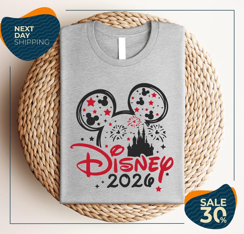 A gray t-shirt with a Disney-themed design, including Mickey Mouse ears and fireworks, is displayed on a woven placemat.