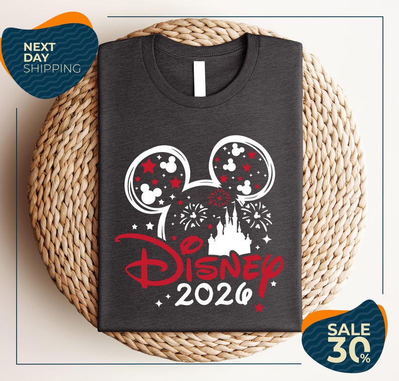 A gray t-shirt with a Disney-themed design, including Mickey Mouse ears and fireworks, is displayed on a woven placemat.