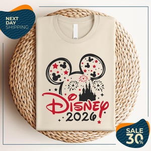 A beige t-shirt with a Disney-themed design, including Mickey Mouse ears and fireworks, is displayed on a woven basket.