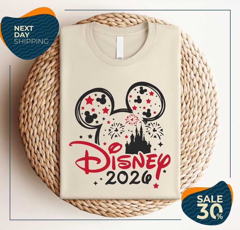 A beige t-shirt with a Disney-themed design, including Mickey Mouse ears and fireworks, is displayed on a woven basket.