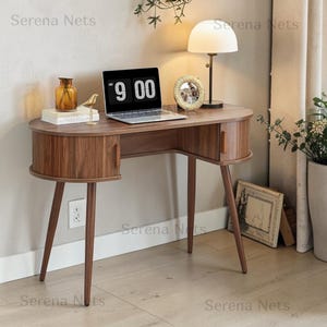 May include: A walnut-finished wooden desk with a curved, oval-shaped top and two rounded side cabinets featuring vertical wooden handles. The desk stands on four tapered legs and holds a laptop displaying 9:00, a lamp, and a small picture frame.
