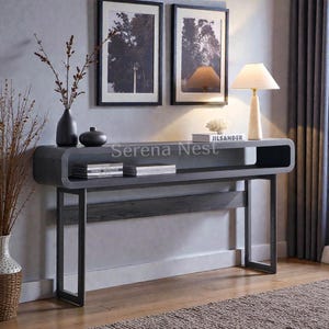 May include: A gray console table with a curved top and a lower shelf. The table has black metal legs and is decorated with vases, books, and a lamp. The text "Serena Nest" is visible on the table.