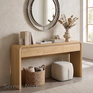 May include: A light oak console table with three drawers, featuring rounded edges. On top sits a round mirror, a white vase with dried branches, books, and a small tray. Below, a woven basket holds two rolled blankets next to a cream ottoman.