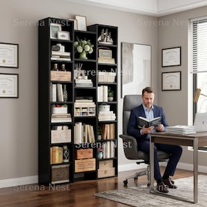 May include: A tall, black bookshelf filled with books, decorative items, and storage boxes. A man in a suit sits in a chair reading a book at a desk. The room has a neutral color palette with framed artwork and certificates.