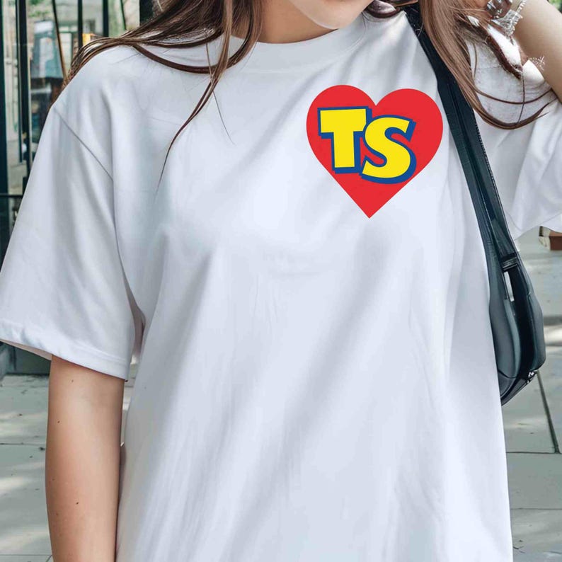 Puede incluir: Una camiseta blanca de manga corta con un gr&aacute;fico de coraz&oacute;n rojo en la parte superior izquierda del pecho. Dentro del coraz&oacute;n rojo, las letras TS est&aacute;n impresas en una fuente amarilla llamativa con un borde azul. Cuello redondo y corte holgado.