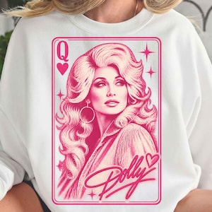 Könnte beinhalten: Ein weißes Sweatshirt mit Rundhalsausschnitt und einem rosa Grafikdruck einer Spielkarte. Das Design zeigt das Porträt einer Frau mit voluminösem Haar, großen Creolen, dem Buchstaben Q und einem Herzsymbol. Unten steht der Schriftzug Dolly.