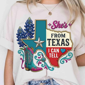 May include: A cream-colored t-shirt featuring a graphic of the state of Texas with a silver star, bluebonnet flowers, and a turquoise and pink cowboy boot. The text reads "She's FROM TEXAS I CAN TELL" with a blue heart icon and decorative swirls.