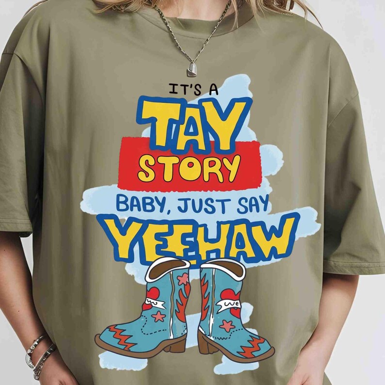 May include: An olive green t-shirt featuring a graphic print with the text IT'S A TAY STORY BABY, JUST SAY YEEHAW in yellow, red, and blue. Below the text are two light blue cowboy boots with red heart and star accents on a light blue background.