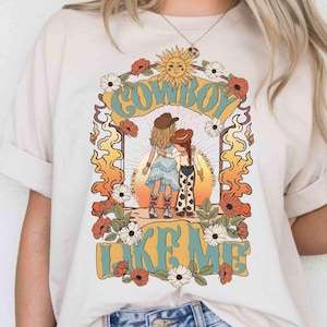 Puede incluir: Una camiseta de color crema con un estampado gr&aacute;fico de dos figuras con sombreros de vaquero de pie juntas. El dise&ntilde;o incluye un sol, flores y llamas, con el texto "COWBOY LIKE ME" en letras grandes y estilizadas de color azul claro y amarillo.