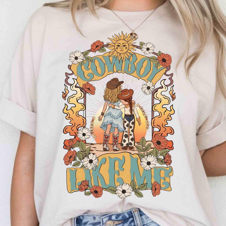 Puede incluir: Una camiseta de color crema con un estampado gr&aacute;fico de dos figuras con sombreros de vaquero de pie juntas. El dise&ntilde;o incluye un sol, flores y llamas, con el texto "COWBOY LIKE ME" en letras grandes y estilizadas de color azul claro y amarillo.