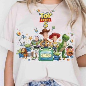 May include: A light beige t-shirt featuring a colorful graphic of Toy Story characters including Woody, Buzz Lightyear, Jessie, Rex, and Mr. Potato Head. The design includes the text "TOY STORY 5" and a green frog-shaped frame with the word "HELLO".