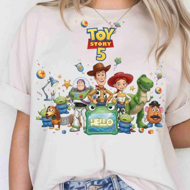 May include: A light beige t-shirt featuring a colorful graphic of Toy Story characters including Woody, Buzz Lightyear, Jessie, Rex, and Mr. Potato Head. The design includes the text "TOY STORY 5" and a green frog-shaped frame with the word "HELLO".