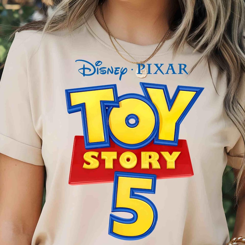 May include: A beige t-shirt featuring a graphic print of the Disney Pixar Toy Story 5 logo. The design includes the blue and yellow Toy Story lettering above a red rectangular background, with the large yellow number 5 positioned below.