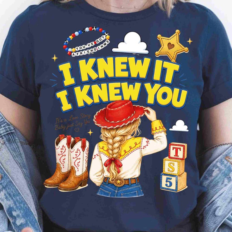 May include: A navy blue t-shirt featuring a graphic of a blonde character with a braid wearing a red cowboy hat, yellow shirt, and blue jeans. The text reads I KNEW IT I KNEW YOU, with illustrations of cowboy boots, a star badge, and toy blocks.