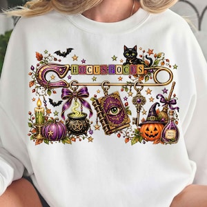 May include: A white sweatshirt featuring a graphic print of a large gold safety pin adorned with the text HOCUS POCUS, a black cat, a purple book with an eye, a cauldron, a pumpkin, a candle, a key, a broom, and a potion bottle with stars.