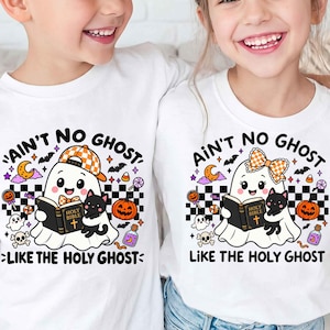 Könnte beinhalten: Zwei weiße T-Shirts mit einer Grafik eines Geistes, der eine schwarze Katze und ein Buch mit dem Titel Holy Bible hält. Der Text lautet AIN'T NO GHOST LIKE THE HOLY GHOST. Das Design enthält Kürbisse, Fledermäuse, Süßigkeiten und ein Schachbrettmuster.