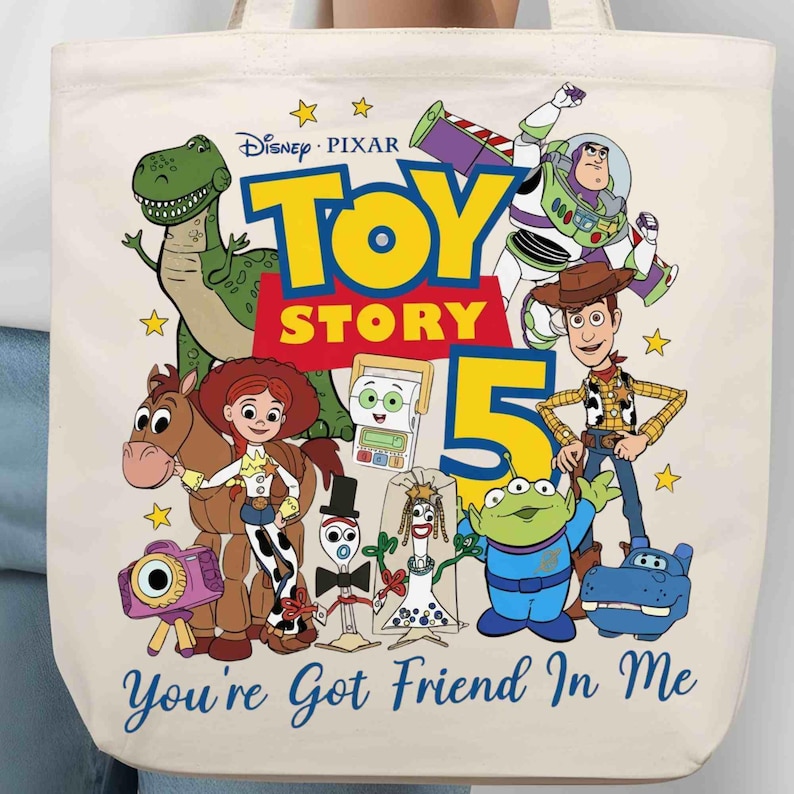 May include: A cream-colored tote bag featuring a colorful Toy Story 5 graphic with characters including Woody, Buzz Lightyear, Jessie, Rex, and others. The text reads Disney Pixar Toy Story 5 and You've Got Friend In Me in blue and yellow.