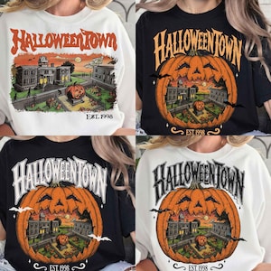 Könnte beinhalten: Eine Kollektion von vier T-Shirts mit einem Grafikdruck einer Stadtszene mit einem großen geschnitzten Kürbis, Fledermäusen und dem Text HALLOWEENTOWN EST. 1998. Die T-Shirts sind in Weiß und Schwarz erhältlich und zeigen detaillierte Gebäude.