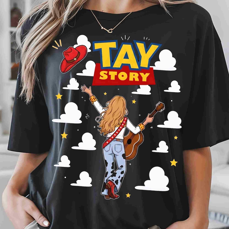 May include: A black t-shirt featuring a graphic print with the text TAY STORY in yellow and red. The design includes a person with long blonde hair playing an acoustic guitar, wearing cow-print trousers, a red hat, and white clouds on a black background.