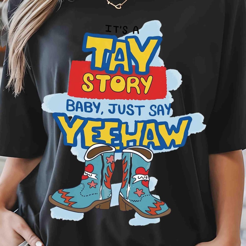 May include: A black t-shirt featuring a graphic with the text IT'S A TAY STORY BABY, JUST SAY YEEHAW in yellow, red, and blue. Below the text are two light blue cowboy boots with red heart and star accents on a light blue brushstroke background.