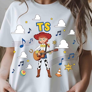Puede incluir: Una camiseta blanca con el gr&aacute;fico de una vaquera de dibujos animados con pelo rojo tocando una guitarra ac&uacute;stica. El dise&ntilde;o incluye el texto TS, notas musicales azules, nubes blancas, estrellas amarillas, una herradura y una estrella.