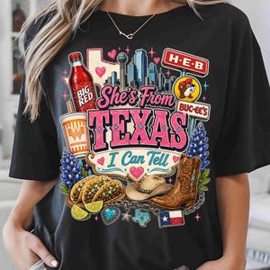 May include: A black t-shirt featuring a graphic print with the text "She's From TEXAS I Can Tell". The design includes a Big Red bottle, a Whataburger cup, a city skyline, H-E-B and Buc-ee's signs, tacos, a cowboy hat, a boot, and a Texas flag.