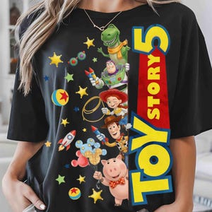 May include: A black short-sleeve t-shirt featuring a graphic print of Toy Story characters including Rex, Buzz Lightyear, Jessie, Woody, and Hamm. The design includes colorful stars, rockets, and balls, with the text Toy Story 5 in large yellow letters.