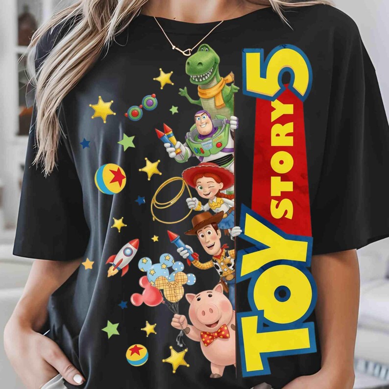 May include: A black short-sleeve t-shirt featuring a graphic print of Toy Story characters including Rex, Buzz Lightyear, Jessie, Woody, and Hamm. The design includes colorful stars, rockets, and balls, with the text Toy Story 5 in large yellow letters.