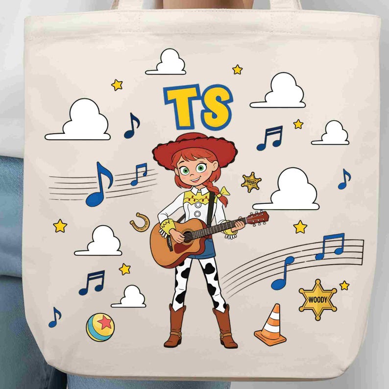 May include: A cream-coloured tote bag featuring a cartoon illustration of a cowgirl character playing an acoustic guitar. The design includes blue musical notes, white clouds, yellow stars, a horseshoe, a ball, a traffic cone, and a sheriff badge with the text Woody. The letters TS are printed in blue and yellow at the top.
