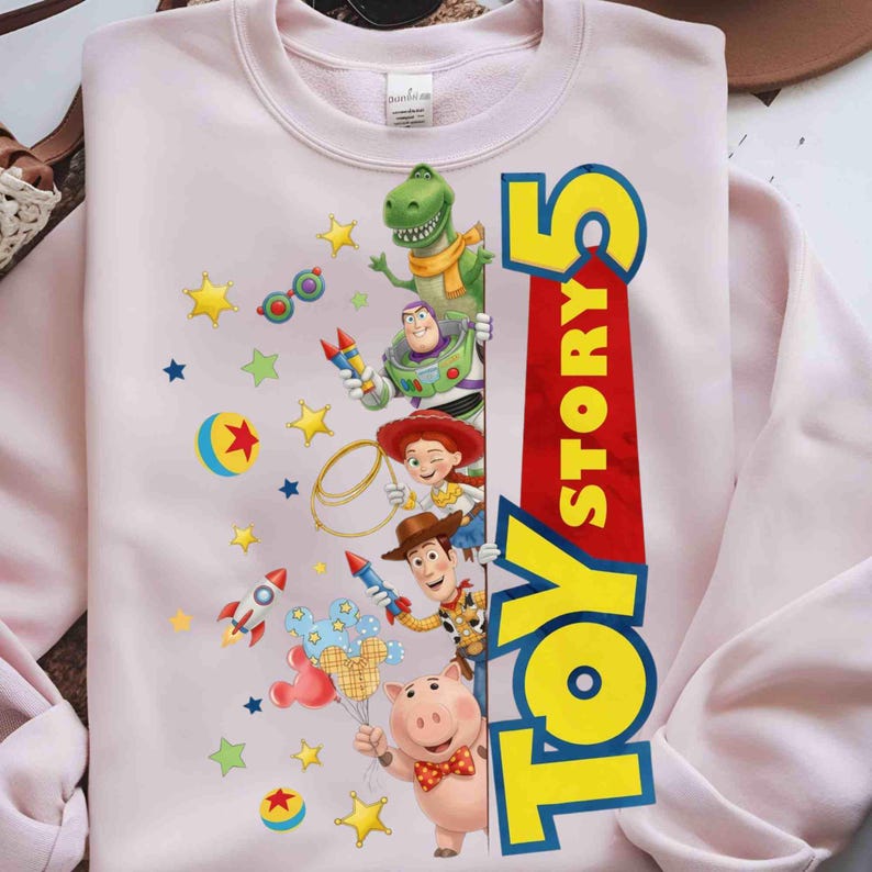 May include: A light pink crewneck sweatshirt featuring a graphic print of Toy Story characters including Rex, Buzz Lightyear, Jessie, Woody, and Hamm, alongside colorful stars, rockets, and balls, with the text Toy Story 5 in large yellow letters.