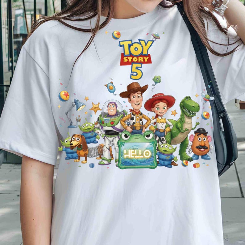 May include: A white short-sleeved t-shirt featuring a colorful graphic print of Toy Story characters including Woody, Buzz Lightyear, Jessie, Rex, Slinky Dog, Mr. Potato Head, and aliens. The text Toy Story 5 and Hello are printed on the front.