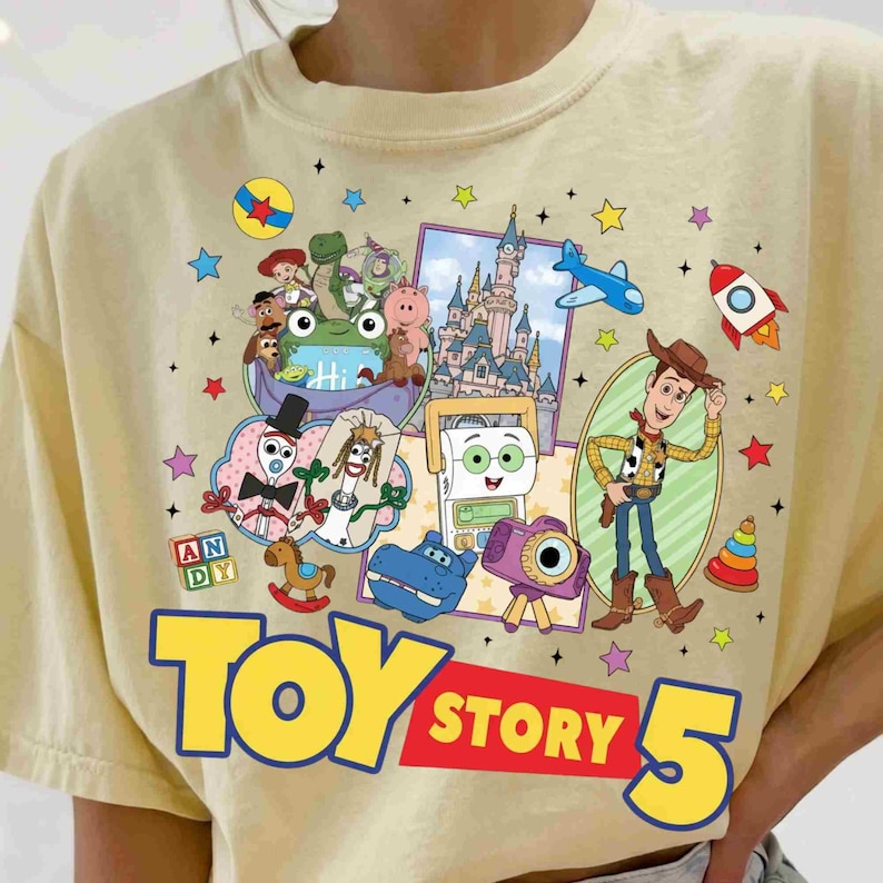 May include: A light yellow t-shirt featuring a colorful graphic print of various animated characters, a castle, a rocket, and a camera. The text "TOY STORY 5" is printed in large yellow and red letters at the bottom, surrounded by small stars.
