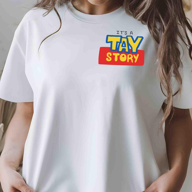 May include: A white short-sleeved t-shirt featuring a graphic on the upper left chest. The graphic displays the text IT'S A TAY STORY in blue and yellow lettering set against a red rectangular background, mimicking a classic movie logo style.