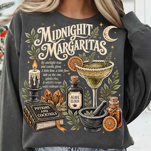 Puede incluir: Una sudadera gris carbón con un estampado gráfico que incluye el texto "Midnight Margaritas". El diseño muestra una vela encendida, una copa de margarita con borde de sal, un libro titulado "Potions and Cocktails", un mortero y cristales.