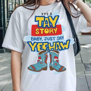 May include: A white short-sleeved t-shirt featuring a graphic print with the text IT'S A TAY STORY BABY, JUST SAY YEEHAW in yellow, red, and blue. Below the text are two light blue cowboy boots with red heart and star accents.