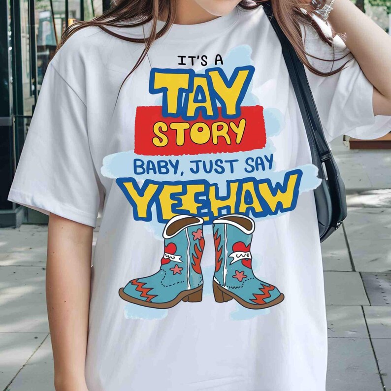 May include: A white short-sleeved t-shirt featuring a graphic print with the text IT'S A TAY STORY BABY, JUST SAY YEEHAW in yellow, red, and blue. Below the text are two light blue cowboy boots with red heart and star accents.